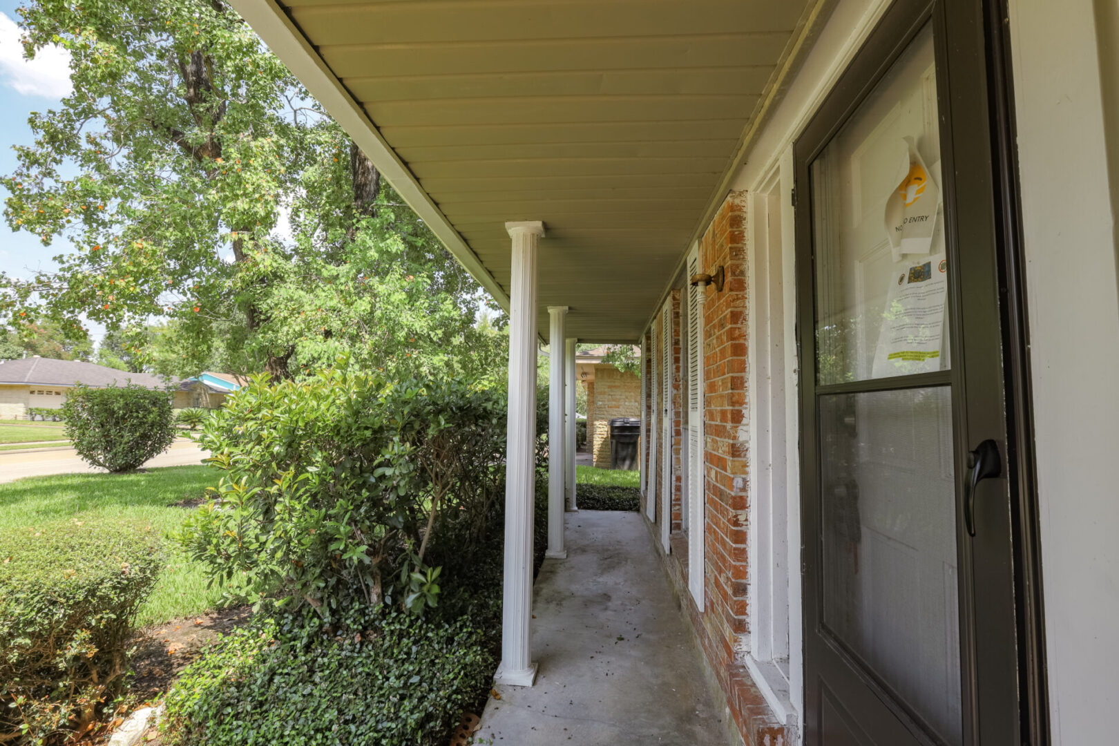 A serene porch with white columns and lush greenery beside a brick house.